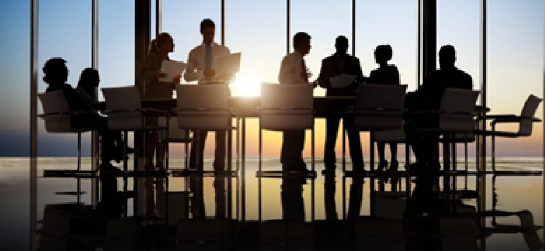 A group of nine silhouetted individuals sits around a table in a modern conference room with a large window. The sun sets in the background, casting a warm glow and creating reflections on the polished floor maintained by commercial cleaning services. Some people hold papers, suggesting a meeting or discussion.