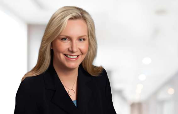 A woman with long blonde hair wearing a black blazer smiles in a brightly lit hallway.