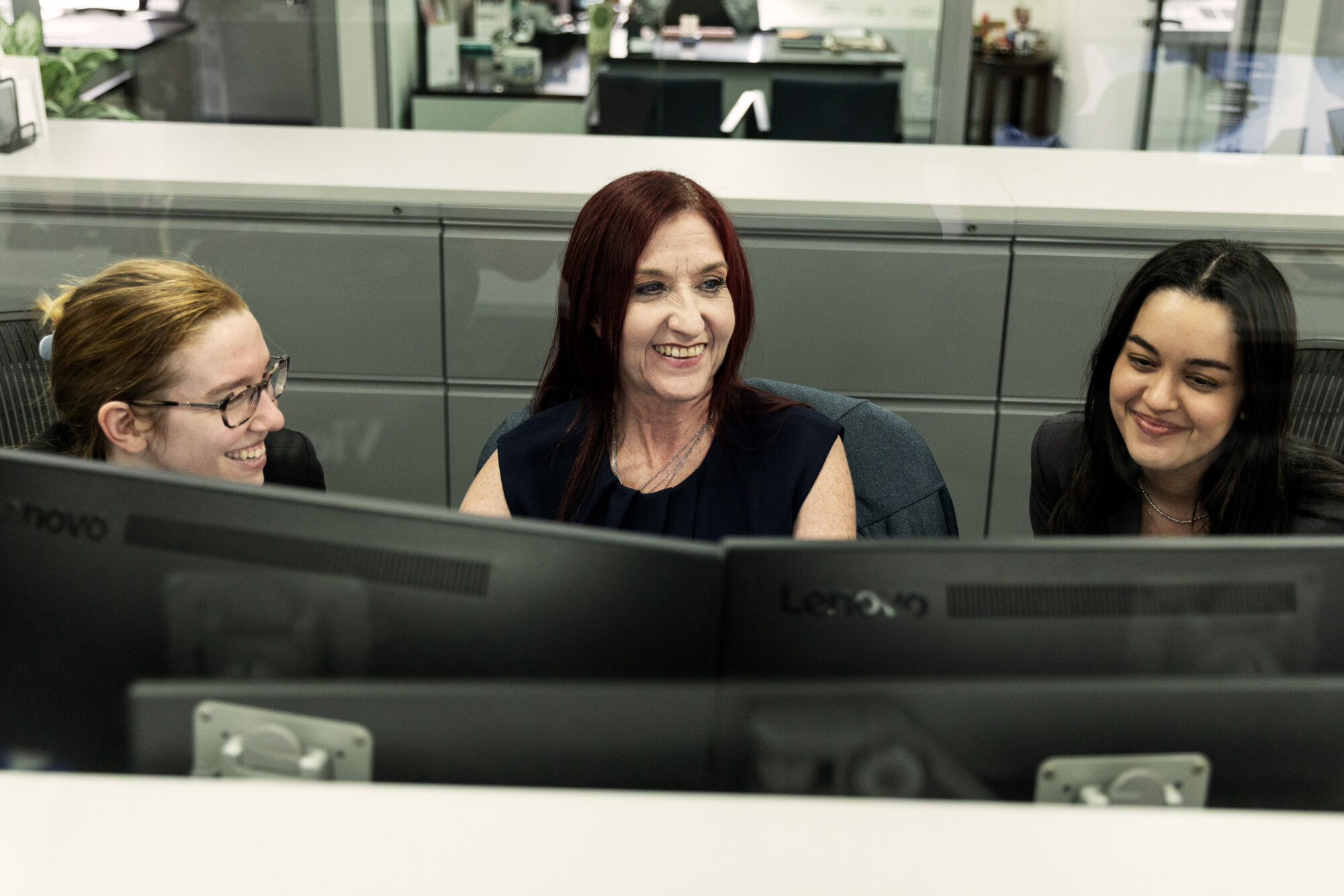 Three women sitting together at desks, smiling, with computer monitors in front of them.