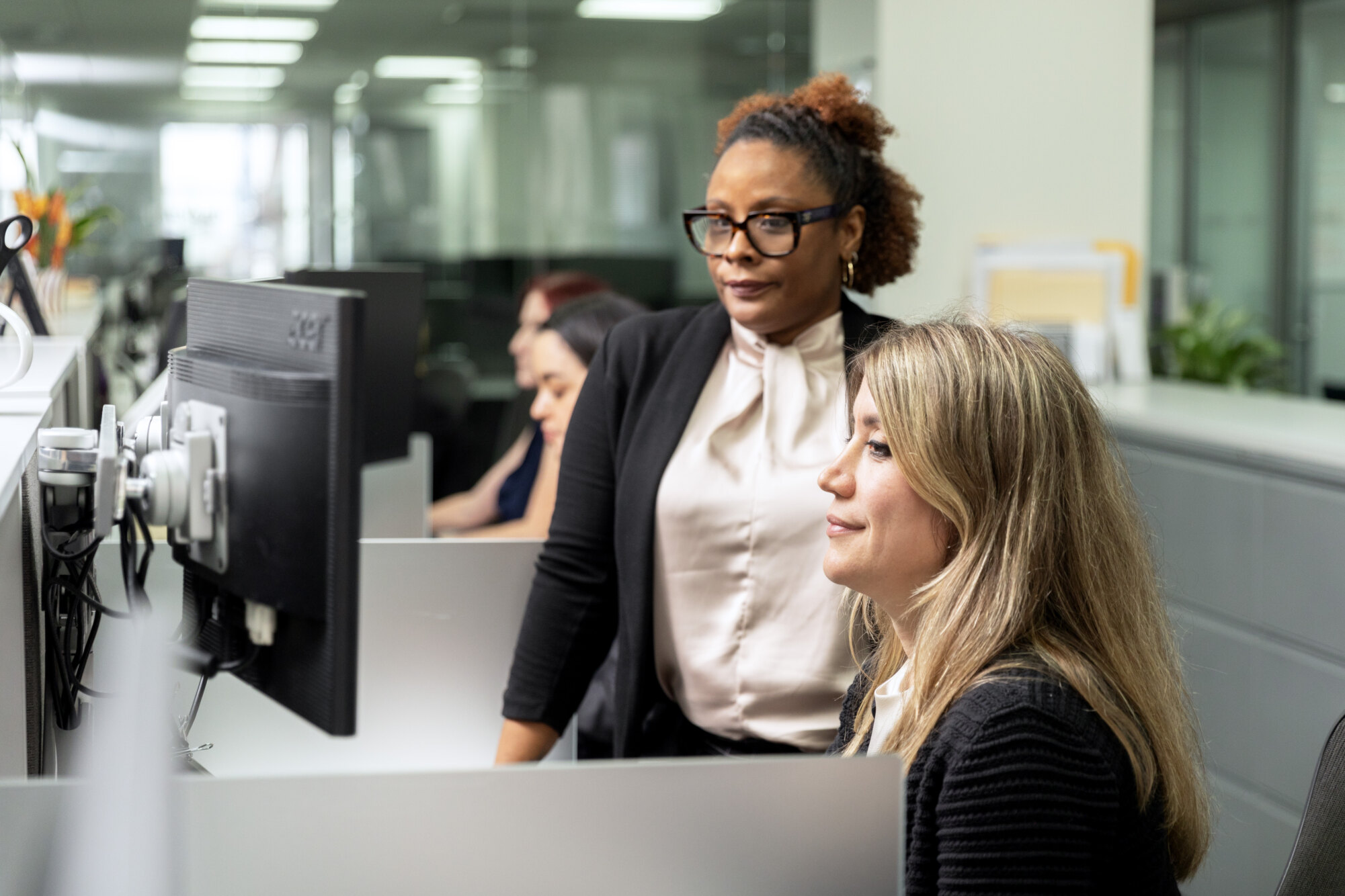 Two women looking at a computer screen in an office setting, with others working in the background.