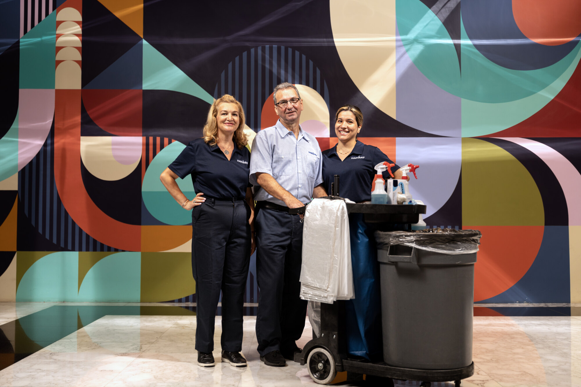 Three cleaning staff stand in front of a colorful abstract wall with a cleaning cart.